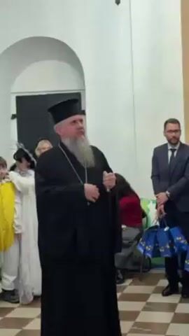An Orthodox priest stands in a room and speaks. He is dressed in a black robe and wears a black hat. He has a gray beard and a silver pendant. He holds a cane in his left hand. He looks to his right as he speaks. There are other people in the background. To the left is a woman in a white dress, and to the right is a man in a suit. There are blue and green gift bags at his feet. The room is white with a checkered tile floor.