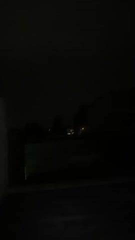 The video shows a dark outdoor scene at night. In the foreground, there is a dark surface, possibly pavement or a floor. To the left, a portion of a brick wall is visible. In the midground, there is a low wall or barrier. Beyond this, the scene opens up to reveal several houses in the distance, with some lights illuminated in their windows or porches. The sky is dark and appears overcast. The overall impression is of a quiet, residential area at night, with minimal light pollution.