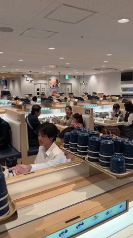 This is a vertical video of a busy sushi restaurant. The camera is positioned at a slightly elevated angle, looking across a conveyor belt system. Rows of booth-style seating are visible, each with a number. Several people are seated in the booths, eating and talking. In the foreground, a young man in a white shirt is looking towards the camera and smiling, with his right arm raised. On the conveyor belt in front of him are several stacks of blue ceramic cups with lids, arranged neatly on wooden trays. The restaurant has bright overhead lighting and a clean, modern aesthetic. The background shows more seating areas and a few framed posters on the wall. The overall atmosphere appears lively and casual.