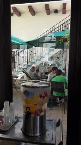 The video shows a person's hands filling a clear plastic cup with fruit-infused water from a large, stainless steel beverage dispenser. The dispenser contains water with slices of oranges and strawberries. The dispenser is on a metal tray with other plastic cups. In the background, through a glass window, is an outdoor courtyard with a staircase adorned with colorful tiles. Several people are seated at tables in the courtyard, one man is reading, and a few others are conversing. The courtyard has umbrellas and potted plants. The overall scene appears to be at a restaurant or cafe with an open-air patio.