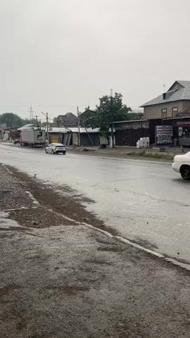 The video shows a street scene on a cloudy day. The road appears wet, suggesting it has been raining or is currently raining. On the left side of the road, a yellow excavator is partially visible, with its bucket lowered to the ground. Further down the road, a white car drives away from the camera, and a black van drives towards the camera. Buildings line the right side of the street, some appearing to be shops or businesses, with a fence or wall behind them. Electrical poles and wires are visible in the background. The overall atmosphere is overcast and somewhat dreary.