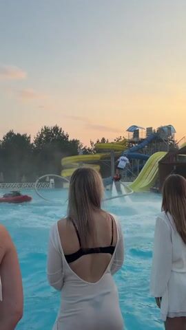 The video opens with a view from behind three people at a water park. In the foreground, the backs of three young women are visible, two in white clothing and one in a white top and black bra. The water in the pool is a bright blue. In the middle ground, a person is water skiing or jet skiing on a red inflatable device, with water spraying around them. In the background, there are water slides, trees, and a sunset sky with pink and orange clouds. The person on the water device is performing a trick, spraying water high into the air. The water park appears to be closing or winding down for the day, given the lighting and the sky.