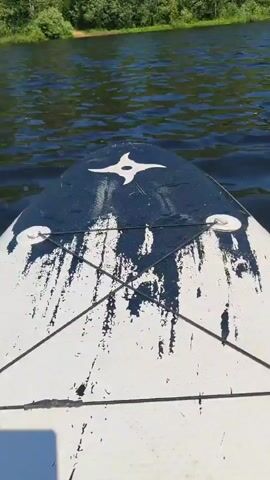 The video shows a first-person perspective of someone stand-up paddleboarding on a body of water, likely a lake or calm river. The view is directed forward, with the front of the paddleboard visible in the foreground. The paddleboard is white with some black markings and a white star symbol on the nose. The water is dark and choppy, with ripples. Trees and lush green foliage line the distant shore. The sun is shining, casting light on the water and the trees. In one shot, another paddleboard is visible to the right of the camera. The overall impression is a peaceful, outdoor recreational activity on a sunny day.