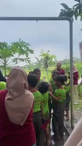 The video shows a group of children covered in mud being washed off under a metal shower structure. The children are wearing matching green t-shirts. Several adults are present, helping and overseeing the children. One adult is wearing a maroon top and a light brown hijab. Another adult wears a black hijab and a black shirt with a logo. The ground is wet and muddy. There is green foliage in the background. The sky appears overcast. One child is seen sitting under a tree. Their face and arms are covered in mud.