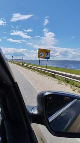 This video appears to be a first-person perspective from inside a car, driving along a road that runs alongside a large body of water, likely a river or lake. The time of day is clearly daytime, with a bright blue sky filled with fluffy white clouds, suggesting fair weather. The road is paved and appears to be in decent condition, with a guardrail separating it from the water's edge. There are some signs visible along the road. One prominent sign is yellow with black text and a red circle containing the number "15" with a symbol of a truck inside, likely indicating a weight limit of 15 tons for trucks. Further down the road, another sign with a red circle and the number "60" is visible, probably a speed limit sign.

The scene is serene and picturesque, with the vast expanse of water stretching to the horizon. The water is a deep blue, reflecting the sky. The vegetation along the roadside is green and appears to be grass and some low-lying plants. A few other vehicles are visible on the road in the distance. The overall impression is one of a leisurely drive through a scenic natural landscape, possibly in a rural or semi-rural area. The reflection in the car's side mirror shows a part of the road and the sky.

The text on the yellow sign is in Cyrillic script. The text above the "15т" reads "ДВИЖЕНИЕ ЛЕГКОВЫХ И ГРУЗОВЫХ АВТОМОБИЛЕЙ". Below that, it says "С МАССАЙ БОЛЕЕ 15 Т." This translates to "MOVEMENT OF PASSENGER AND TRUCK VEHICLES WITH A MASS GREATER THAN 15 T." Therefore, the sign indicates a weight restriction for vehicles. The "60" sign is a standard speed limit sign.

The location context provided is Gorodets, Russia, which aligns with the Cyrillic script on the signs. The gentle movement of the car suggests a smooth journey. There are no people clearly visible within the frame of the car's interior, but the perspective implies the presence of a driver and possibly passengers. The overall mood is peaceful and expansive, indicative of a pleasant travel experience on a beautiful day.