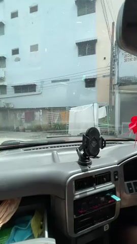 The video appears to be captured from inside a car, looking out through the windshield. In the foreground, the dashboard of the car is visible, with a phone mount attached to it. The phone mount holds a black smartphone. Beyond the windshield, the scene is an alleyway between two buildings. The building on the left is painted in light green and has multiple windows. The building on the right is painted in a pale blue and also has numerous windows and air conditioning units. Laundry is hanging on a line between the buildings, with various colored fabrics visible. There are also signs on the building to the left, one with red and yellow lettering. The overall impression is that of an urban environment, possibly in a warm climate, given the presence of air conditioning units and hanging laundry. The car seems to be stationary or moving very slowly down the alleyway.