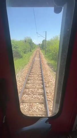The video presents a point-of-view shot from inside a moving train, looking out through the front window down a set of railway tracks. The tracks stretch into the distance under a clear blue sky. Green trees and vegetation line both sides of the tracks. The camera is positioned inside the train cabin, with parts of the red interior visible on either side of the window frame. The motion of the train is evident from the shifting perspective and the rattling sounds. The overhead wires for the electric train are also visible above the tracks.