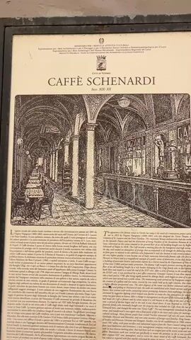 The video begins with a close-up of a framed historical plaque that is mounted on a wall. The plaque has an illustration of what appears to be a coffee shop or cafe, with tables and chairs arranged in a long, narrow room with arched ceilings. Text is present on the plaque, but it is too blurry to read in detail, though some words like "CAFFÈ SCHENARDI" and "SECC. XIX-XX" are discernible. The plaque is mounted at eye level and shows signs of age and wear. The video then pans slightly to the right, showing more of the wall, which is textured and painted a light color, with some peeling paint. The camera then moves to the right, revealing a close-up of a closed metal roll-up door, with a small, square sign featuring the "illy" logo mounted to the wall to its right, next to a bell. The video then pans further right and upwards, showing a building facade with a large arched entryway, also covered by a closed metal roll-up door. Above the door, the name "SCHENARDI" is displayed in large, gold-colored letters. Further to the right, another arched entryway is visible, also covered by a roll-up door. The building's facade is a muted, peachy-orange color, with some areas of exposed brick or plaster. The overall scene depicts the exterior of a historical building, likely a cafe, which appears to be closed for business. The lighting suggests it is daytime.
