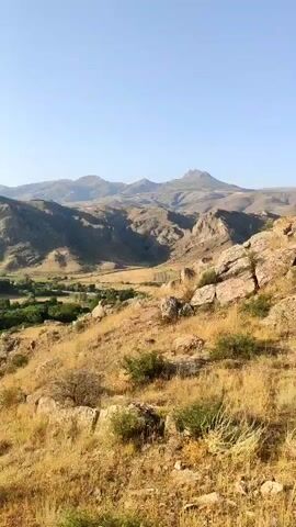 A landscape video opens with a panoramic view of a valley nestled between rocky hills and mountains. The foreground features dry, yellowed grass and scattered rocks, with sparse bushes. As the camera pans, a village comes into view, characterized by houses with reddish-brown roofs and lush green trees. The village is situated in the valley floor, surrounded by verdant vegetation. Beyond the village, rugged, light-brown mountains rise, their slopes etched with shadows. The sky above is a clear, pale blue. The lighting suggests it is daytime, with the sun casting shadows and illuminating the terrain. There are no people or discernible animals visible in the video. The overall impression is of a remote, natural landscape with a small settlement.