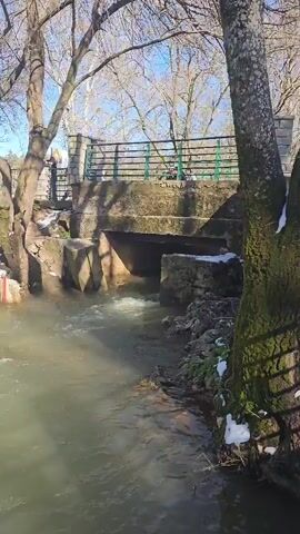 The video shows a river flowing through a park in winter. The water is a light green color and moves quickly under a stone bridge. The banks of the river are lined with trees and patches of snow. There are a few people walking on the other side of the river.