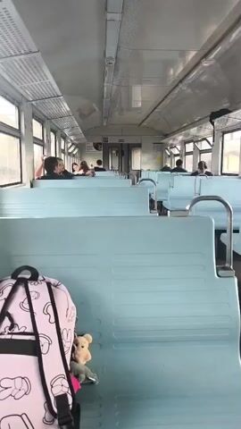 The video shows the interior of a train carriage. Light blue seats are visible on both sides of the aisle, some of which are occupied by passengers. In the foreground on the left, a pink backpack with a Mickey Mouse-like pattern is placed on a seat. A small stuffed bear is attached to the backpack. The train appears to be moving, as suggested by the slight blur in the background and the subtle sway that can be inferred from the motion of the camera. The overall lighting is natural, coming from the windows on the side.