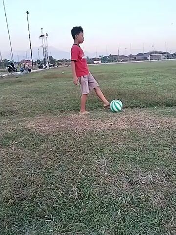 This media file captures a young boy playing soccer in an outdoor field. He is barefoot and wearing a red and white striped t-shirt and light-colored shorts. The boy is actively engaged with a soccer ball, which has green and white stripes.

The scene appears to be set in a park or sports field with a grassy ground. In the background, there are trees, some buildings, and what looks like streetlights or poles. The sky is clear and suggests it is either early morning or late afternoon, with the soft light of dusk or dawn. The overall atmosphere seems peaceful and recreational. There is another person in the distance to the left, and another boy in yellow shorts further to the right, also on the field, hinting at a casual game or practice session. The setting of Dari, Indonesia, provides a geographical context for this scene of outdoor recreation.