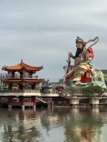 The video shows a large statue of a seated figure holding a sword in a body of water, with a traditional pagoda structure next to it. The statue is brightly colored, with a long beard and a crown. The pagoda has a multi-tiered roof and red columns. The water reflects both the statue and the pagoda. Birds fly in the sky, which is overcast. The buildings can be seen in the distance.