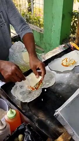 A man wearing a gray t-shirt is preparing food on a flat-top grill. He is standing behind the grill and is in the process of assembling what appears to be a wrap. He is holding a piece of white cheese in his left hand and is about to place it on top of some filling on a thin white wrapper. Another wrap is already on the grill with filling and a slice of cheese on top, as well as a dollop of red sauce. To the left of the grill, there is a green bucket containing shredded white material, possibly cabbage or cheese, and a plastic bag with more shredded material. There are also several condiment bottles, a small black bag, and what look like small eggs in a container. The grill is on a wooden cart, and behind the cart is a green wall with a fence and some greenery visible beyond. The lighting suggests it is daytime.