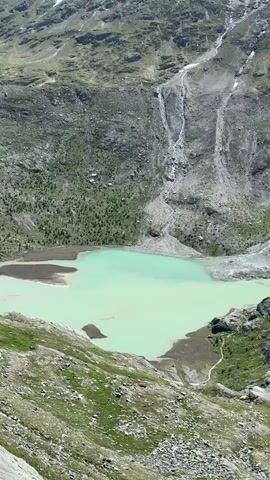 This is a vertical video of a mountain lake and a mountain slope. The lake is a bright turquoise color and is surrounded by rocky terrain and some greenery. Waterfalls cascade down the mountain slope, which is covered in rocks, sparse vegetation, and patches of snow. The sky is partly cloudy. The camera appears to be stationary and is looking down at the lake and mountain.