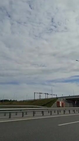 A car drives under an overpass on a cloudy day. The camera is mounted on a vehicle driving on a highway. The car is white. The highway has a metal guardrail. The sky is full of white clouds. There are streetlights on the highway.