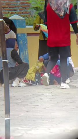 Four people, three of whom are wearing hijabs, are gathered outdoors on a paved surface. One woman in a blue hijab and grey top is squatting. Another woman in a purple hijab and blue top is also squatting and appears to be interacting with a young child. In front of them are several water bottles. A third woman in a yellow top and black hijab is standing and holding colorful fabric. A fourth person, wearing a red top and black pants, is standing behind them, facing away from the camera. A young girl in a red t-shirt and blue jeans is standing to the left and facing away from the camera. The background shows a brick wall and a low blue and yellow structure. A metal pole is in the foreground.