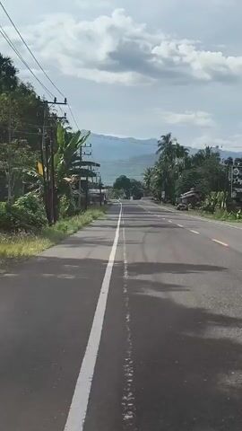 The video shows a straight, paved road stretching towards a mountain range in the distance. The road is marked with a solid white line in the center. Trees and vegetation line both sides of the road. Utility poles and wires run along the left side. There are scattered buildings along the roadside. The sky is partly cloudy. A motorcycle drives in the distance along the road.