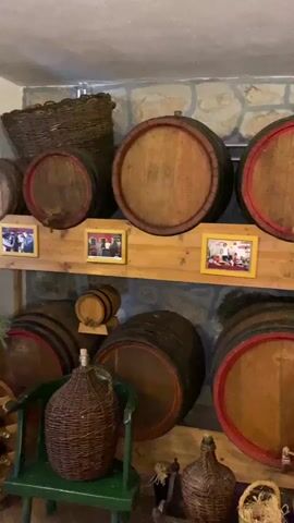 The video shows an indoor scene, likely a cellar or storage room, filled with numerous wooden barrels of various sizes. Most of the barrels feature red painted bands around their ends, and some have spigots. They are arranged on two levels of wooden shelving and also placed on the floor. Between the barrels on the top shelf, several small, framed pictures, likely photographs, are displayed.

Other objects in the room include a large woven wicker basket on the top left, and several wicker-covered glass bottles (demijohns) on the floor, some with green wooden stands. A person wearing a blue short-sleeved shirt is partially visible on the right side of the frame, moving their right arm. The walls behind the barrels are constructed from light-colored stone blocks, suggesting a rustic or traditional setting. Some copper and wooden bowls are also visible on the shelves. The audio contains voices speaking, indicating conversation. The overall impression is that of a traditional storage area, possibly for wine or other beverages, in Meterizi, Montenegro.