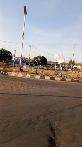 The camera moves forward to capture a street-level view of a paved road. A grassy area enclosed by a metal fence is visible to the right. Within the fenced area, there are multiple white metal structures of unknown purpose. Behind the fenced area, a gas station and other buildings are visible. The sky is bright blue, suggesting a sunny day. There is a sign with a large white outline shape and white lettering on top of the grassy area, but the lettering is difficult to read.