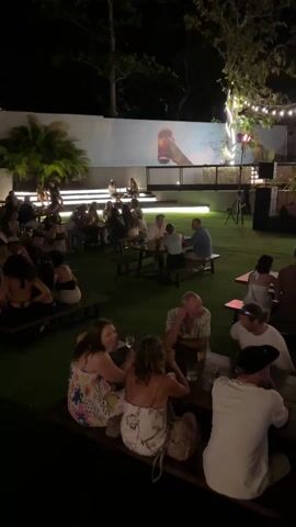 The video shows a lively outdoor event at night. Numerous people are seated at picnic tables, chatting and enjoying themselves. The area is covered in artificial turf, and there is a large screen projecting an image, possibly a movie or advertisement, on a white wall at the back. There's an elevated seating area at the rear. The scene includes lush greenery and string lights, contributing to a relaxed atmosphere. The camera pans from right to left, capturing the breadth of the gathering and the diversity of the crowd. A two-story building with a glass-enclosed upper level can be seen along the left side of the frame. Servers can be seen moving around, seemingly attending to the guests.
