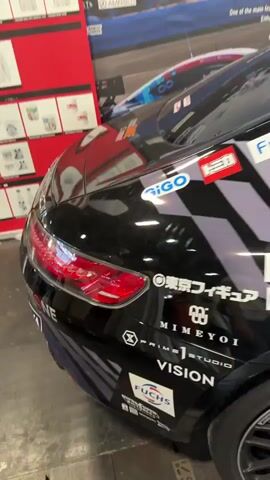 The video pans across the side of a black car with numerous sponsors' logos and graphics. The graphics include a checkered pattern in purple and light grey, and some teal and light blue abstract designs. Some of the sponsors' logos visible are "VISION," "WINSTON," "CHRONO-CARS," "KYB," and "YAMAHA." The perspective then shifts to the side of a white car covered in anime-style graphics, featuring a character with turquoise hair and headphones, possibly Hatsune Miku. Various sponsor logos are also visible on this car, including "TAITO," "Digi Club," and "AMBOSS FOREST." People are visible in the background of the white car's shot, walking around an indoor event space, possibly a convention or exhibition. The lighting suggests an indoor environment.
