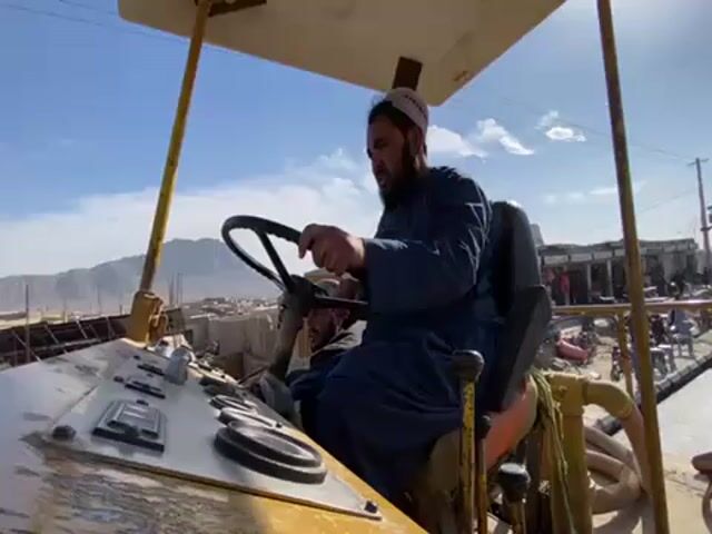 The video shows a man in a blue robe and a white skullcap operating a piece of heavy construction equipment, possibly a road roller. He has a beard and is focused on driving. Another person is sitting next to him, partially visible. The machinery is yellow with a simple canopy structure. The control panel is visible, displaying several gauges. The surrounding environment appears to be an open construction site in a town with mountains in the distance. Several people are working in the background, along with construction materials and equipment. The sky is clear and blue.