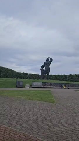 This video showcases a large, dark-colored monument dedicated to war heroes. The central subject is a colossal statue of a muscular man, depicted in a heroic pose with his right arm raised to his forehead as if surveying the distance. He holds a large sword vertically in his left hand, resting its base on the ground. The statue stands on a grassy mound, backed by a dense line of dark green trees.

In front of the statue, there is a long, dark-colored memorial wall. The wall is divided into sections, with rows of names inscribed on plaques. A prominent inscription on a central plaque reads "НИКТО НЕ ЗАБЫТ НИЧТО НЕ ЗАБЫТО" which translates to "No one is forgotten, nothing is forgotten." Red flowers are visible on some of the plaques, suggesting recent or ongoing remembrance.

The foreground is paved with reddish-brown brickwork. The overall setting appears to be a solemn memorial park. The sky is overcast with a muted gray, indicating a cloudy day, possibly late afternoon or early morning given the ambient light. The mood conveyed is one of respect, remembrance, and national pride.

The location is identified as Свіслацкі сельскі Савет, Belarus, a rural council area. The monument likely commemorates fallen soldiers or victims of war, serving as a significant landmark for remembrance and historical reflection in the region.
