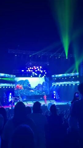 A low-angle shot shows a concert crowd with their backs to the camera, watching a singer on stage. The performer, dressed in a red suit, stands in the center of the stage, singing into a microphone. Behind him, a large screen displays a tropical scene of mountains and waterfalls. Additional performers are arranged on stage around him. Bright green spotlights shine down from above, creating a dynamic lighting effect.  In the background, a disco ball hangs from the ceiling, adding to the concert atmosphere.