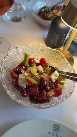 The video shows a close-up of a Greek salad in a clear, textured bowl. The salad contains cherry tomatoes, feta cheese, olives, cucumbers, and red onions. A spoon and fork are resting in the salad. In the background, another bowl of salad is visible, along with a bottle of olive oil and a small lamp with a gold shade. A glass of pink liquid is also visible on the left side of the frame. The setting appears to be a restaurant with a white tablecloth. The camera slowly zooms out slightly, revealing more of the table and the surrounding environment.