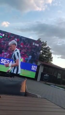 The video starts with a close-up of a large outdoor screen displaying a soccer match, with the logo of "S NOVA" visible. The screen shows a player in a black and white striped jersey. The word "SAME" is displayed at the bottom of the screen. A woven chair is visible in the foreground. The camera then pans upwards, showing a single-story building with large windows. The camera continues to tilt up, revealing a picturesque sky with shades of blue, gray, and orange, indicating either sunrise or sunset. Trees, including a palm tree, are silhouetted against the bright horizon. A soccer goal can be seen in the background, implying a backyard setting.