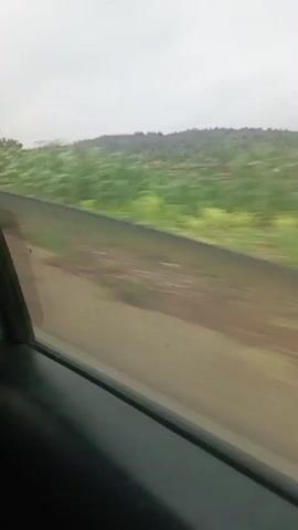 The video shows a view from inside a vehicle traveling along a dirt road. The road is brown and dusty, with patches of reddish-brown soil. To the left of the road, there is a low, dark-colored barrier. Beyond the barrier, there is a strip of green vegetation, and further in the distance, rolling hills covered in green foliage. The sky is overcast and gray. In the foreground, the interior of the vehicle is visible, including a dark-colored door panel and a portion of the window frame. As the vehicle moves, buildings and trees become visible in the distance. The buildings are white with red roofs. A gas station with a red canopy can be seen at one point. The motion blur indicates the vehicle is in motion. There are no people clearly visible in the video. The environment appears to be rural or semi-rural.