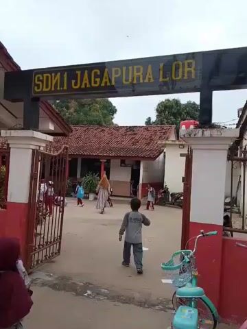 The video shows the entrance to SDN 1 JAGAPURA LOR, a school, on a cloudy day.  A black sign with yellow lettering identifies the school.  An open gate leads into a schoolyard where several children and at least one adult are walking.  The school buildings have reddish-brown tiled roofs and white walls.  A turquoise bicycle is parked near the gate.  Some people wear school uniforms.  One child wearing a maroon outfit walks directly toward the camera through the gate.