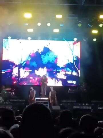 The video shows a live performance on a stage. A woman in a long dress sings into a microphone, flanked by two men in sleeveless tops who perform choreographed movements. Behind them is a large screen displaying a stylized, colorful animated landscape with trees, bushes, and a watery path. Stage lighting illuminates the performers, and a large crowd is visible in the foreground.