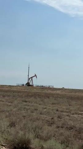 A lone oil pump jack stands in a flat, arid landscape under a clear sky with wispy clouds. The pump jack is enclosed by a fence and appears to be in operation, with its arm moving up and down. The surrounding terrain is covered in dry grass and sparse vegetation. The video is shot from a distance, giving a wide view of the scene.