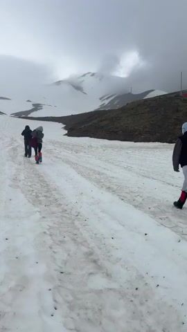 The video shows two people walking away from the camera on a snowy, mountainous path. The path is somewhat muddy and has tire tracks. The sky is overcast and the mountains in the background are covered in snow and fog. The person in the lead appears to be wearing a dark jacket and pants, and carrying a backpack. The person behind them is wearing a red and blue backpack and appears to be wearing colorful pants. The environment is rugged and appears to be at a high altitude.