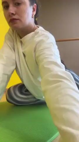 A woman is doing a yoga stretch on a green yoga mat. She is wearing a white long-sleeved shirt and gray and white tie-dye leggings, as well as white socks. She has her hair pulled back and is wearing earrings. She is in a room with a yellow wall and a taupe wall, and there is a barre attached to the wall. She begins in a seated position on top of a gray and white striped pillow, and then moves into a lying position to do the stretch.