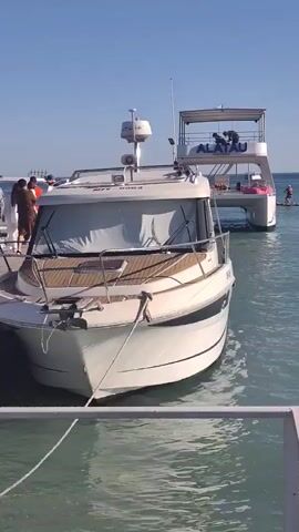 This video captures a sunny day at a harbor in Aktau, Kazakhstan. In the foreground, a white motor yacht is docked, its bow facing the viewer. The yacht has a wooden deck area and a covered cabin with white curtains obscuring the interior. A blue and white rope secures it to the dock.

Behind the motor yacht, a larger catamaran-style boat is also docked. This boat has multiple levels and a canopy-style top deck. Several people are visible on the upper deck of the catamaran. The word "ALATAU" is clearly visible in blue lettering on the side of the catamaran.

The water is a calm, light blue-green, suggesting fair weather. The sky is clear and a bright blue, indicating a sunny day, likely late morning or early afternoon given the brightness and the shadows. In the distance, several other boats and what appear to be buoys or markers are visible on the horizon. The overall atmosphere appears peaceful and recreational, with people enjoying time on the water.