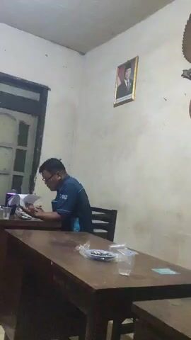A man wearing a dark blue polo shirt with a light blue logo is sitting at a wooden desk. He is wearing glasses and appears to be reading some papers. In front of him on the desk are some plastic cups and a small plate with what looks like snacks. Behind him, on the wall, is a framed picture of a man in a suit. To his left is a window with wooden shutters. The room appears to be an office or classroom.