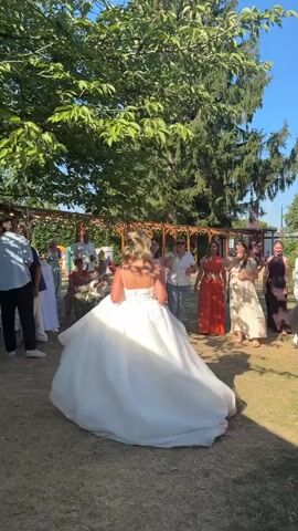 A wedding reception is taking place outdoors on a sunny day. A bride in a white wedding dress is about to throw her bouquet. She is facing away from the camera, her back to the guests, who are gathered in a semi-circle facing her. The guests are dressed in various attire, and some appear to be holding their phones up to capture the moment. The bride is holding a bouquet of flowers in her right hand. The background shows trees, a wooden pergola with lights, and a clear blue sky. The ground is covered in grass and gravel.