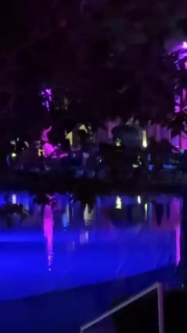 The video displays a nighttime scene, possibly at a resort, featuring a swimming pool illuminated with vibrant blue and pink lights. The pool is surrounded by tropical foliage and structures, including white cabanas with draped fabric. In the background, a building is lit with pink lights. The camera pans across the scene, capturing the reflections of the lights on the water and the ambient atmosphere. There are no people visible in the video.