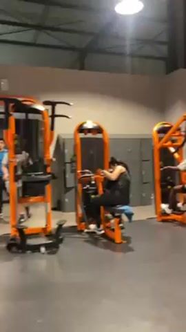In a spacious, well-lit gym, several individuals are engaged in exercising on stationary machines with distinctive orange frames. The gym features a high ceiling supported by visible steel beams and a row of gray lockers lining one wall. A woman in a black workout outfit is seated, operating one of the machines. A younger person in a white shirt is also using a machine, while another person with a white visor stands nearby.  Another man in a blue shirt and patterned pants appears to be interacting with the woman on the machine, possibly providing guidance. The camera pans slowly, capturing more of the gym's interior and the people within it.