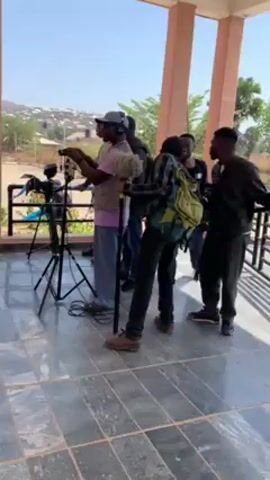 A group of people are gathered on a tiled porch, likely filming a scene. In the foreground, a video camera is set up on a black tripod. A person wearing headphones is adjusting equipment attached to the camera. Several other people stand nearby, some holding boom microphones and equipment. A young woman walks into the frame, heading towards the camera setup. Behind them, there is a view of a sunny landscape with buildings and hills. The setting appears to be outdoors, next to a building with peach-colored pillars.