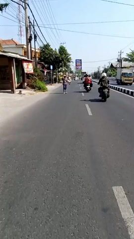 The video shows a street scene from the perspective of someone moving forward on a vehicle. Several people are visible, including two individuals on motorcycles traveling in the same direction as the camera, and several pedestrians walking on the side of the road. There is a yellow truck parked on the left side of the road, along with some construction materials. The environment appears to be an urban or semi-urban area with buildings, trees, and power lines lining the street. The day is sunny.