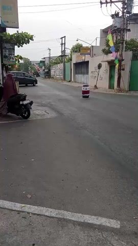 A street scene in an urban environment. A paved road runs through the center of the frame, with buildings and trees lining the sides. A few motorbikes and a car are present on the road. A person is riding a motorbike, and another person is riding a motorbike carrying a cylindrical object. A person is standing near a motorbike. There is a traffic cone in the middle of the road. The sky is overcast.