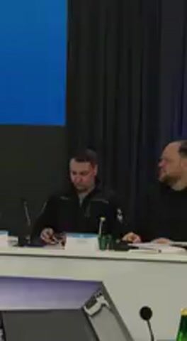 The video shows an indoor scene, likely a conference or meeting room, in Barvinok, Ukraine. Two men are seated at a long table. In the foreground, a man with short dark hair, wearing a dark jacket over a lighter shirt, is visible from the waist up. He is seated at the table, looking intently to his left, out of the frame, possibly at a speaker. His expression is serious and attentive. In front of him on the table is a white nameplate or card, the text on which is not clearly legible. A microphone is positioned near him, and a green bottle (possibly water or a soft drink) is also on the table. A portion of a laptop or tablet is visible to his left. To his right, another man is partially visible, also seated at the table. Behind the men, there is a large, solid blue screen or display on the left side, and dark curtains on the right. The overall impression is that of a formal gathering or press event.