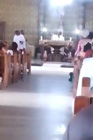 The video shows an interior view of a church during a service. Rows of wooden pews are visible on either side of a central aisle, with attendees seated. Two individuals in white robes stand to the left, near the front. At the altar, a person in white clerical attire is conducting the service. Above the altar, a crucifix is mounted on the wall. The scene is brightly lit from overhead, and the general ambiance is one of reverence and traditional church service.