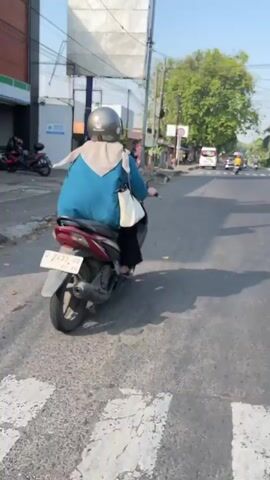 A person wearing a blue poncho and a silver helmet rides a scooter down a paved road. The scooter is dark in color with a license plate that has red lettering. The person is carrying a white bag on their left shoulder. The road is lined with buildings and trees. The sky is clear and blue. There are other vehicles and people in the background.