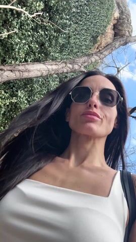 A woman with long, dark hair is featured in a selfie-style video, appearing to walk outdoors on a sunny day. She is wearing dark, polygonal-shaped sunglasses with a silver frame and a white top with wide straps and a square neckline. A tattoo, resembling a crescent moon and possibly a star or sun, is visible on her right shoulder.

The video begins with a close-up shot of her face, angled slightly upwards, capturing her in motion as she walks. Her lips are slightly parted in a soft, pleasant expression. In the initial frames, dense green foliage and some tree branches are visible behind her. As the video progresses, she continues to walk, and the camera follows her movement. The background shifts to reveal more bare tree trunks and branches, suggesting a wooded area. Towards the end of the clip, a dirt or gravel path becomes visible behind her, confirming she is on a trail or walking path. The sky, visible in glimpses through the trees, is blue with some clouds. Her hair moves slightly with her steps, and she maintains a consistent, calm facial expression throughout the short video.