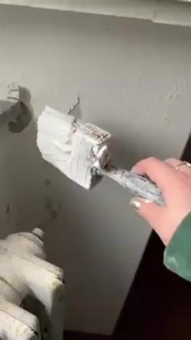 A hand wearing a green sleeve is shown painting a white wall with a white brush. The wall has some cracks and imperfections. A pipe and a radiator are visible to the left of the area being painted. The floor is dark.